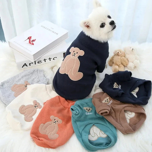 Pets-Shop. A white dog wearing a navy blue sweater with a bear design, surrounded by similar sweaters in different colors. Shopets-Amazon.